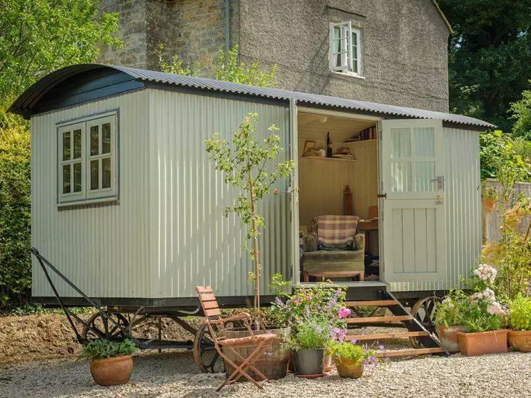 Plankbridge Cabin in a garden setting surrounded by beautiful potted plants and through the open door a comfy armchair is visible.